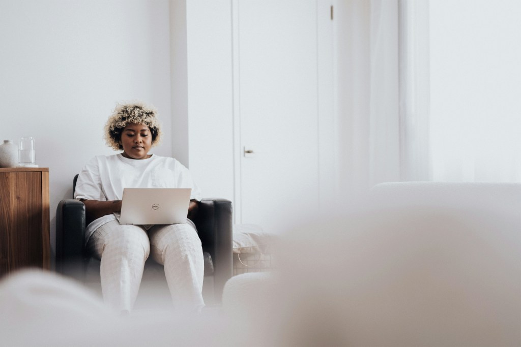 woman with dark skin and multi-tones hair is wearing a white tee and using a Dell laptop while sitting in a dark leather chair in a bright, white room.