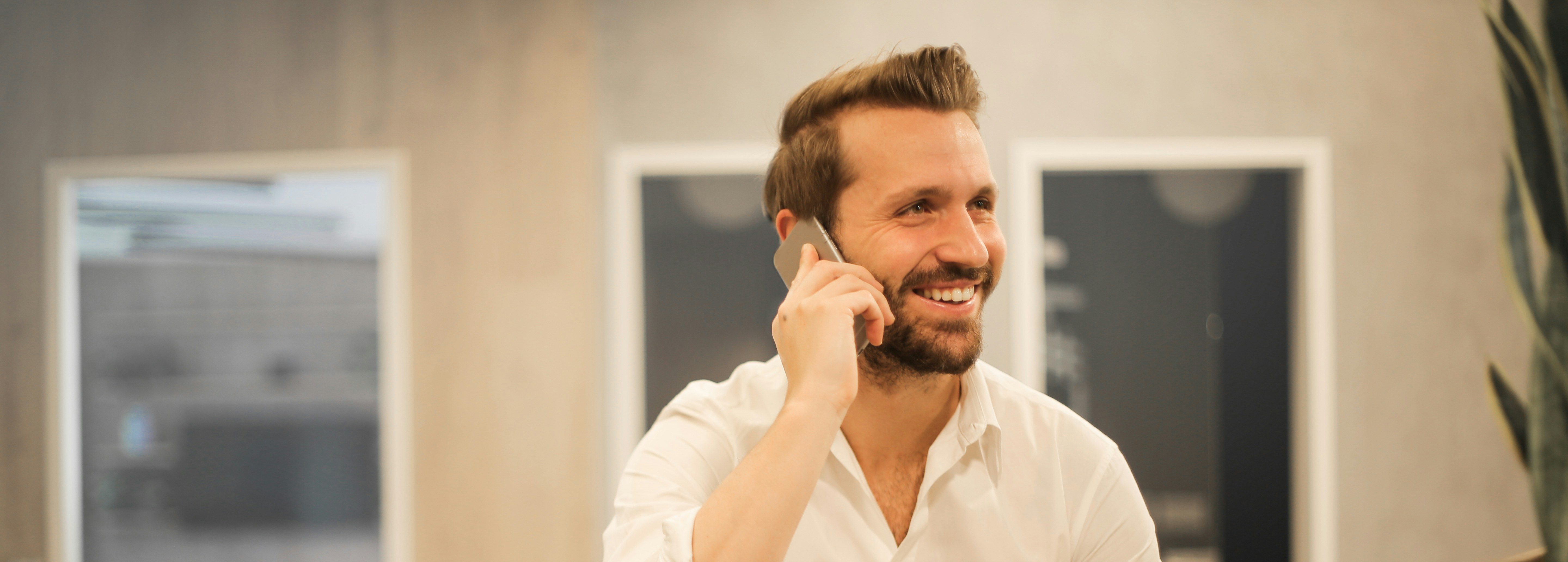 gentleman with short brown hair and a beard talking on a cell phone in an office with his laptop open in front of him. He is smiling and wearing a crisp, white dress shirt.